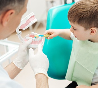 Little boy in dental chair brushing model teeth held by dentist